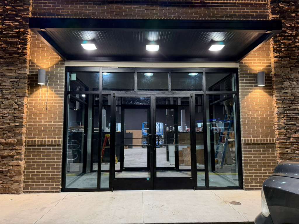 Frontal view of a brick storefront entrance with black aluminum doors and canopy lighting